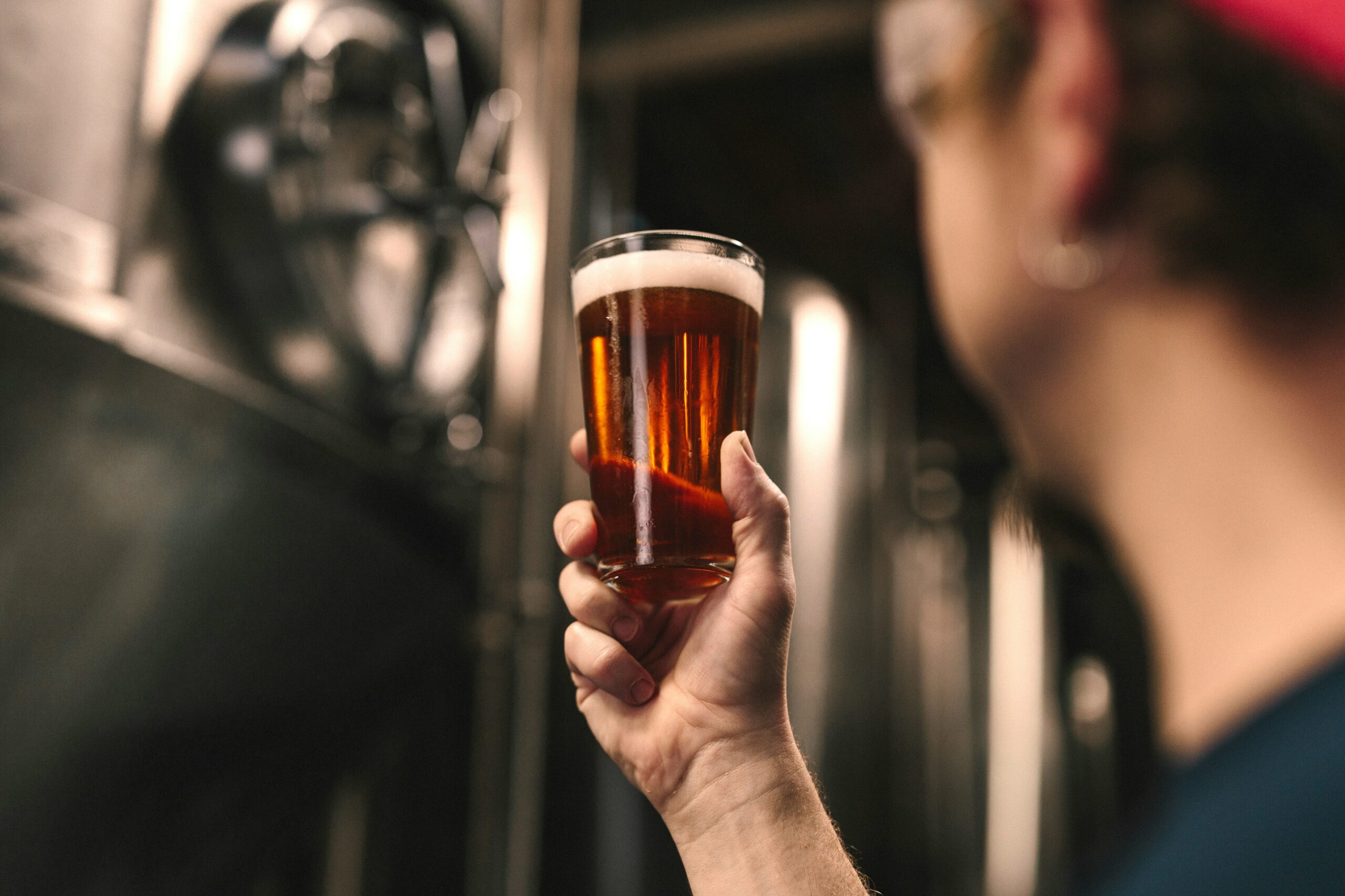man holding glass of beer with brewing equipment in the background
